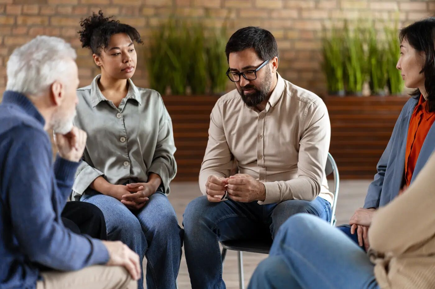 A diverse group of adults sitting in a circle, leaning in and listening to one another in a warm, sunlit room